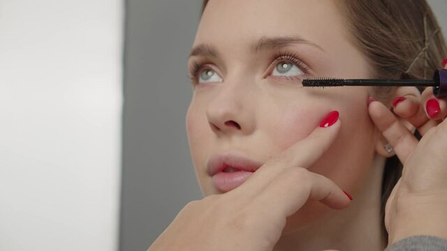 Make-up artist in photo studio doing makeup for beautiful girl. Woman making up using comb for eyelashes. Young woman making makeup in dressing makeup room. Making mua. Applying mascara to the lashes