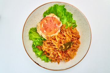 Stewed white cabbage with carrots, one piece of fried chicken fillet, one piece of round tomato, one bay leaf and two lettuce leaves on a white round plate on a white background.