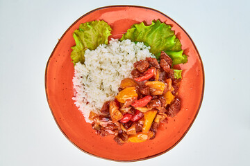 Boiled white rice porridge with meat stew with red, yellow sweet pepper and lettuce leaves in a round orange plate on a white background.
