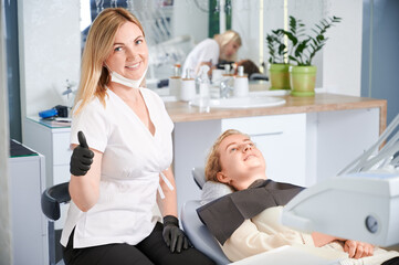 Cheerful female stomatologist showing approval gesture and smiling while woman lying in dental chair. Joyful dentist in sterile gloves doing thumbs up while sitting beside patient in dental office.