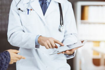 Unknown red-bearded doctor and patient-woman discussing current health examination while sitting and using tablet computer in sunny clinic, close-up. Medicine concept