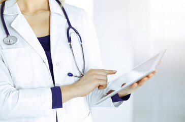 Unknown woman-doctor is holding a tablet computer in her hands, while standing in a sunny clinic cabinet. Female physician at work, close-up. Perfect medical service in a hospital. Medicine concept