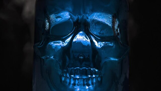 Glass Skull Glowing with Blue Light. A bright, rich amethyst-blue vessel in the form of a glass skull, from which fumes emanate and slowly descend along the edges against a black background