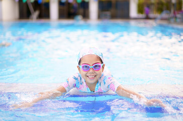 Asian child cute or kid girl wearing swimming suit and goggles on edge swimming pool and smile with happy fun in water park for learn and enjoy training swim or refreshing to exercise on summer school