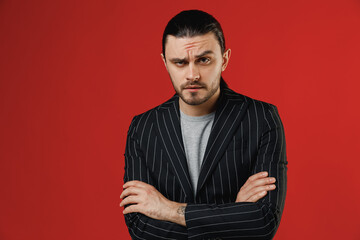 Fototapeta premium Young serious shrewd confident strict businessman latin man in black striped jacket grey shirt hold hands crossed folded isolated on red background studio studio portrait. Concept of people lifestyle.