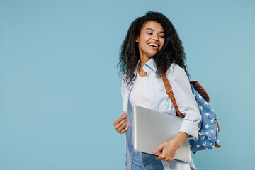 Obraz premium Young smiling african american girl teen student in denim clothes backpack hold closed laptop pc computer look aside isolated on blue background Education in high school university college concept.