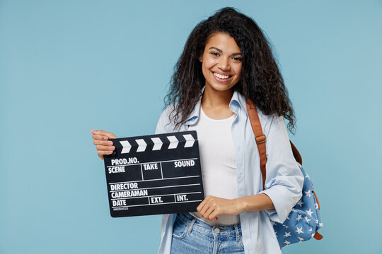 Young fun african american girl teen student wear denim clothes backpack holding classic black film making clapperboard isolated on blue background Education in high school university college concept.