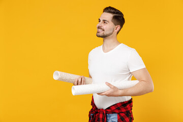 Young smiling employee handyman man wear t-shirt holding wallpaper rolls look aside isolated on...