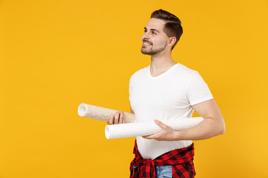 Young smiling employee handyman man wear t-shirt holding wallpaper rolls look aside isolated on yellow background studio. Instruments accessories for renovation apartment room. Repair home concept.