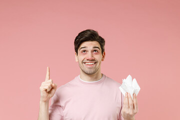 Smiling happy sick ill allergic man has red eyes runny nose suffer from allergy hold paper napkin pointing finger above on workspace promo advert area isolated on pastel pink color background studio. - obrazy, fototapety, plakaty