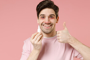 Sick unhealthy ill allergic man has red watery eyes runny stuffy sore nose suffer from allergy symptoms hay fever hold bottle uses instill nasal drops isolated on pastel pink color background studio.