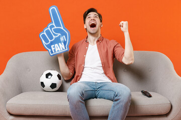Young excite man football fan in shirt foam glove finger support favorite team with soccer ball sit...