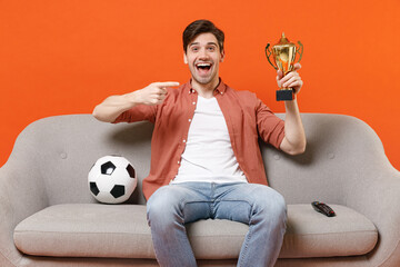 Young excited man football fan in shirt support team with soccer ball sit home sofa watch tv live...