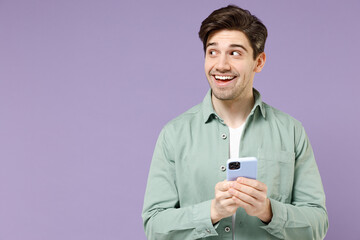 Young pensive dreamful caucasian man in mint shirt white t-shirt use mobile cell phone chat online in social network look aside isolated on purple background studio portrait. People lifestyle concept