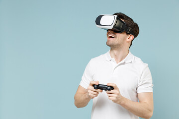 Young gambling man 20s in white casual basic t-shirt play pc game with joystick console watch in vr headset pc gadget isolated on pastel blue color background studio portrait People lifestyle concept