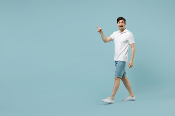 Full length young fun caucasian man wear white basic t-shirt point finger aside on workspace area copy space mock up isolated on pastel blue color background studio portrait. People lifestyle concept