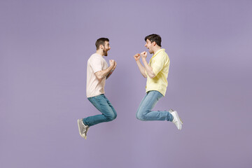 Fototapeta na wymiar Side view full length two young happy men friends together in casual t-shirt looking to each other do winner gesture clench fist jump high isolated on purple background studio People lifestyle concept