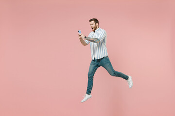 Fototapeta na wymiar Full length young overjoyed unshaven man 20s wear blue striped shirt white t-shirt jump high hold mobile cell phone chat online isolated on pastel pink background studio portrait Tattoo translate fun