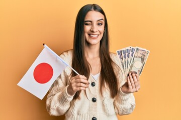Beautiful brunette young woman holding japan flag and yen banknotes winking looking at the camera...