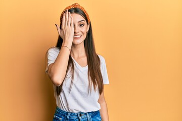 Beautiful brunette young woman wearing casual white t shirt covering one eye with hand, confident smile on face and surprise emotion.