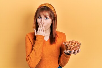 Redhead young woman holding peanuts covering mouth with hand, shocked and afraid for mistake....