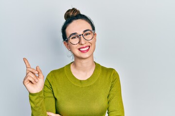 Young hispanic girl wearing casual clothes and glasses with a big smile on face, pointing with hand and finger to the side looking at the camera.