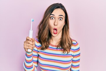 Young hispanic girl holding electric toothbrush scared and amazed with open mouth for surprise, disbelief face