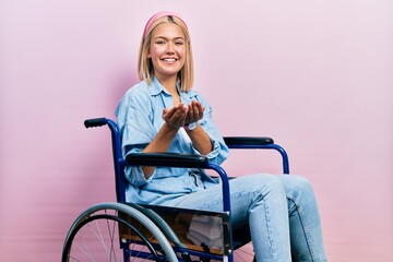 Beautiful blonde woman sitting on wheelchair smiling with hands palms together receiving or giving...