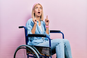 Beautiful blonde woman sitting on wheelchair amazed and surprised looking up and pointing with...