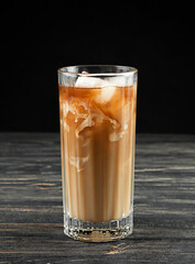 Iced coffee with ice and milk in a tall glass on a black wooden background.