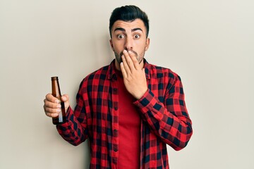 Young hispanic man drinking a bottle of beer covering mouth with hand, shocked and afraid for...