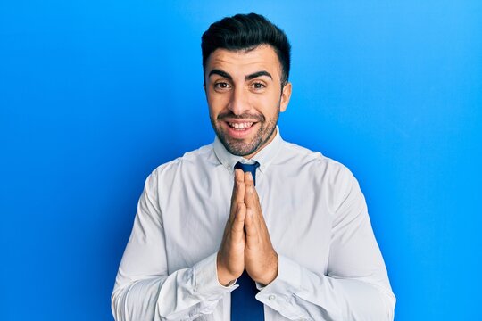 Young hispanic man wearing business clothes praying with hands together asking for forgiveness smiling confident.