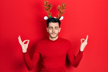Young hispanic man wearing cute christmas reindeer horns relax and smiling with eyes closed doing meditation gesture with fingers. yoga concept.