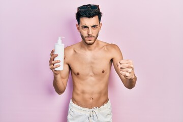 Fototapeta premium Young hispanic man wearing swimsuit and holding sunscreen lotion annoyed and frustrated shouting with anger, yelling crazy with anger and hand raised