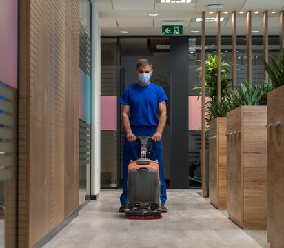 Cleaner with the mask on his face cleans the hard floor in the office with walk behind scrubber dryer machine.Cleaning and maintenance concept