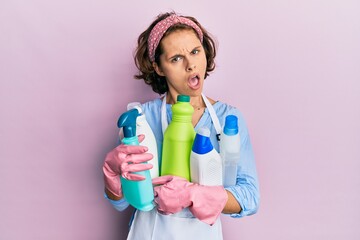 Young brunette woman wearing cleaner apron holding cleaning products in shock face, looking skeptical and sarcastic, surprised with open mouth