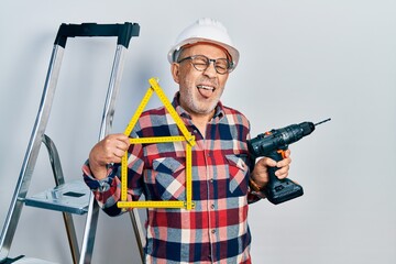 Handsome mature handyman holding screwdriver wearing hardhat by construction stairs sticking tongue out happy with funny expression.