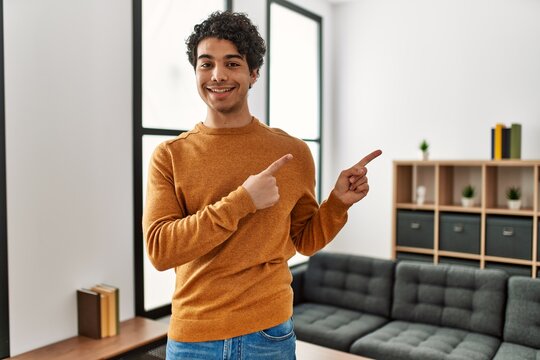 Young Hispanic Man Wearing Casual Clothes Standing At Home Smiling And Looking At The Camera Pointing With Two Hands And Fingers To The Side.