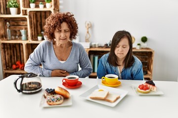 Family of mother and down syndrome daughter sitting at home eating breakfast with hand on stomach because nausea, painful disease feeling unwell. ache concept.
