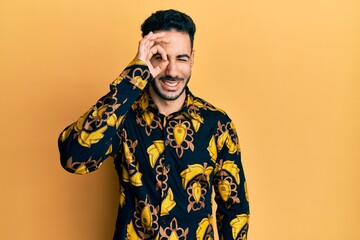 Young hispanic man wearing casual shirt doing ok gesture with hand smiling, eye looking through fingers with happy face.