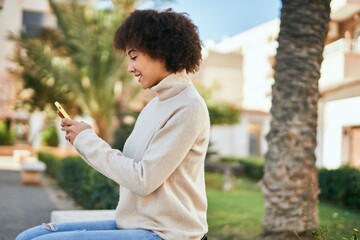 Young hispanic girl smiling happy using smartphone sitting on the bench at the park.