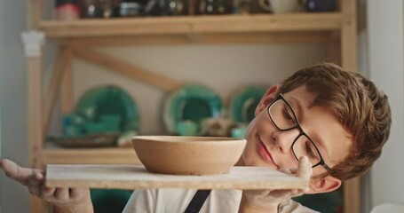 Young teen boy with glasses looks at his product a plate of clay portrait looks at the camera smiling standing in a pottery workshop