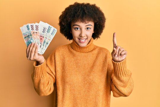 Young hispanic girl holding czech koruna banknotes smiling with an idea or question pointing finger with happy face, number one