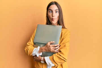 Fototapeta premium Young beautiful woman holding laptop making fish face with mouth and squinting eyes, crazy and comical.