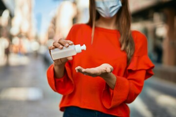 Fototapeta premium Young hispanic woman wearing medical mask and using hand sanitizer gel at the city.