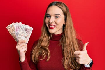 Young blonde woman holding turkish lira banknotes smiling happy and positive, thumb up doing excellent and approval sign