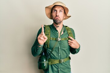 Handsome man with beard wearing explorer hat and backpack pointing up looking sad and upset,...