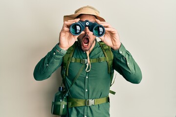 Handsome man with beard wearing explorer hat looking through binoculars afraid and shocked with...