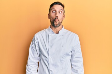 Handsome man with beard wearing professional cook uniform making fish face with lips, crazy and comical gesture. funny expression.