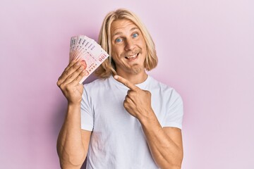 Caucasian young man with long hair holding 100 new taiwan dollars banknotes smiling happy pointing with hand and finger